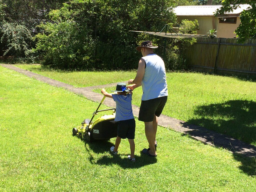 Man and child pushing a lawn mower across a sunny suburban backyard lawn