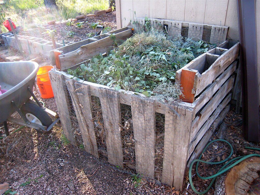 Wooden pallet compost bin heaped with green garden waste beside a wheelbarrow